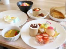 Flat lay of assorted desserts on white plates: strawberry cake, macarons, fruit tart, peaches, and a cup with paw-print cocoa.