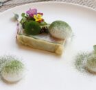 Fine dining plate: a rectangular layered dish topped with greens and edible flowers, with two white quenelles dusted in green powder on a white plate.