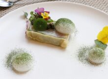 Fine dining plate: a rectangular layered dish topped with greens and edible flowers, with two white quenelles dusted in green powder on a white plate.