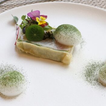 Fine dining plate: a rectangular layered dish topped with greens and edible flowers, with two white quenelles dusted in green powder on a white plate.