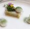 Fine dining plate: a rectangular layered dish topped with greens and edible flowers, with two white quenelles dusted in green powder on a white plate.