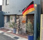 German flag hoisted on a pole outside a blue stucco building with stone veneer and potted plants