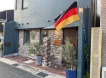 German flag hoisted on a pole outside a blue stucco building with stone veneer and potted plants
