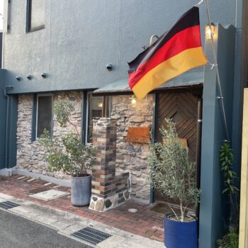 German flag hoisted on a pole outside a blue stucco building with stone veneer and potted plants