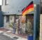 German flag hoisted on a pole outside a blue stucco building with stone veneer and potted plants
