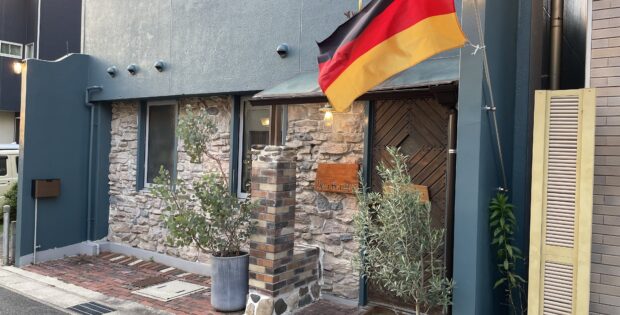 German flag hoisted on a pole outside a blue stucco building with stone veneer and potted plants