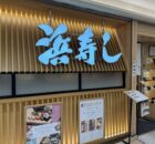 Exterior view of a Japanese restaurant with a large bright blue kanji sign mounted on a vertical wooden slat wall and takeout menus posted nearby.