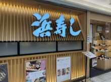 Exterior view of a Japanese restaurant with a large bright blue kanji sign mounted on a vertical wooden slat wall and takeout menus posted nearby.