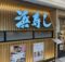 Exterior view of a Japanese restaurant with a large bright blue kanji sign mounted on a vertical wooden slat wall and takeout menus posted nearby.