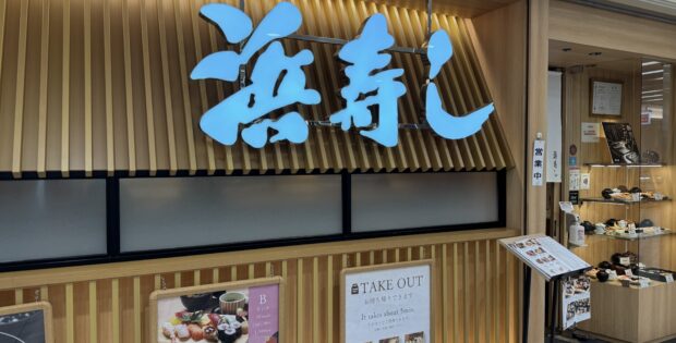 Exterior view of a Japanese restaurant with a large bright blue kanji sign mounted on a vertical wooden slat wall and takeout menus posted nearby.