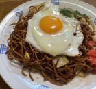 Plate of fried noodles with a sunny-side-up egg on top, garnished with red pickled ginger and greens on a decorative white dish.