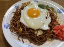 Plate of fried noodles with a sunny-side-up egg on top, garnished with red pickled ginger and greens on a decorative white dish.