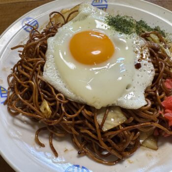 Plate of fried noodles with a sunny-side-up egg on top, garnished with red pickled ginger and greens on a decorative white dish.