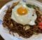 Plate of fried noodles with a sunny-side-up egg on top, garnished with red pickled ginger and greens on a decorative white dish.