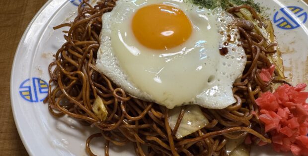Plate of fried noodles with a sunny-side-up egg on top, garnished with red pickled ginger and greens on a decorative white dish.