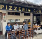 Outdoor seating area of Pizzeria Conza with three men at a long wooden table looking at menus outside the restaurant signage in view.