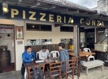 Outdoor seating area of Pizzeria Conza with three men at a long wooden table looking at menus outside the restaurant signage in view.