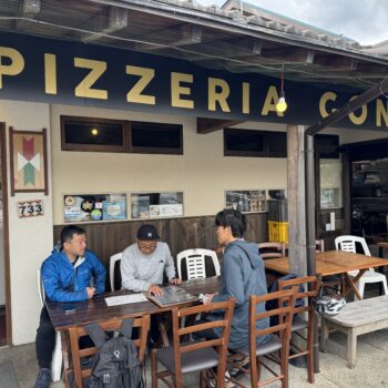 Outdoor seating area of Pizzeria Conza with three men at a long wooden table looking at menus outside the restaurant signage in view.