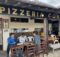 Outdoor seating area of Pizzeria Conza with three men at a long wooden table looking at menus outside the restaurant signage in view.