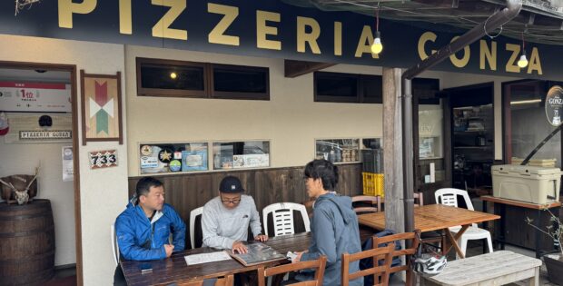 Outdoor seating area of Pizzeria Conza with three men at a long wooden table looking at menus outside the restaurant signage in view.