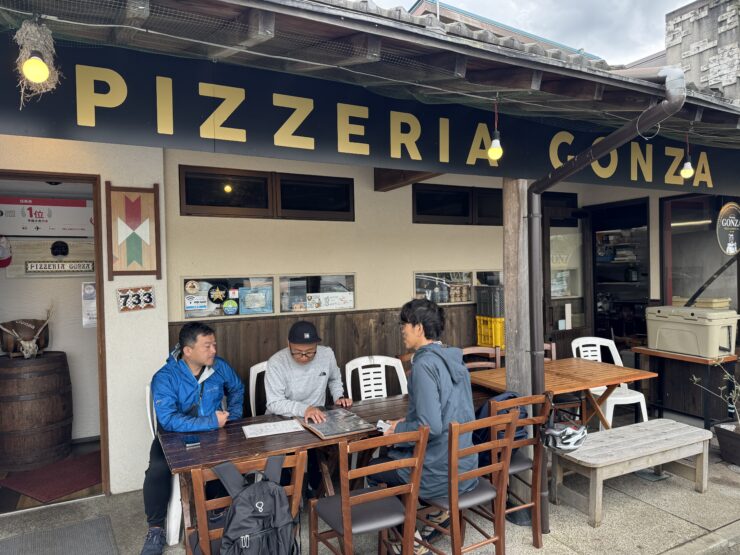 Outdoor seating area of Pizzeria Conza with three men at a long wooden table looking at menus outside the restaurant signage in view.