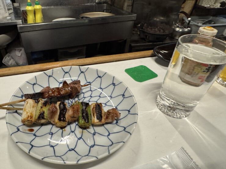 Skewered grilled meat and vegetables on a blue patterned plate with sauce drips, in a kitchen setting with a glass of water nearby.