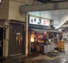 Nighttime street scene with a Japanese food stall, red lanterns, and illuminated signboards outside a narrow shop under an awning.