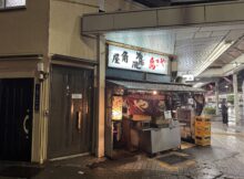 Nighttime street scene with a Japanese food stall, red lanterns, and illuminated signboards outside a narrow shop under an awning.
