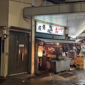 Nighttime street scene with a Japanese food stall, red lanterns, and illuminated signboards outside a narrow shop under an awning.