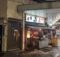 Nighttime street scene with a Japanese food stall, red lanterns, and illuminated signboards outside a narrow shop under an awning.
