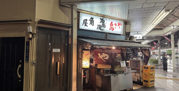 Nighttime street scene with a Japanese food stall, red lanterns, and illuminated signboards outside a narrow shop under an awning.