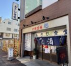 storefront of a Japanese eatery with blue noren curtains and bold red/blue kanji on a beige sign above the entrance, brick exterior on a sunny street