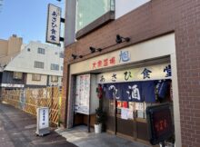 storefront of a Japanese eatery with blue noren curtains and bold red/blue kanji on a beige sign above the entrance, brick exterior on a sunny street