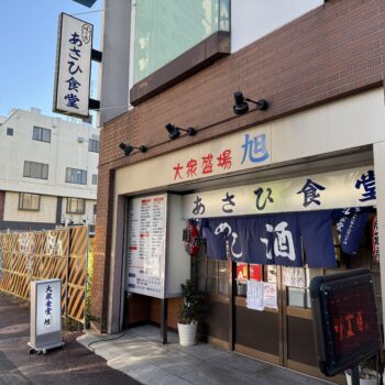 storefront of a Japanese eatery with blue noren curtains and bold red/blue kanji on a beige sign above the entrance, brick exterior on a sunny street