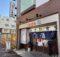 storefront of a Japanese eatery with blue noren curtains and bold red/blue kanji on a beige sign above the entrance, brick exterior on a sunny street