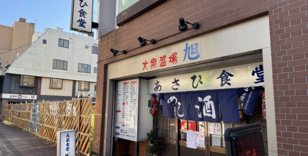 storefront of a Japanese eatery with blue noren curtains and bold red/blue kanji on a beige sign above the entrance, brick exterior on a sunny street