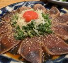 Thinly sliced seared tuna sashimi on a blue plate, sprinkled with sesame seeds and shredded onions and greens, topped with a cherry tomato.