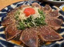 Thinly sliced seared tuna sashimi on a blue plate, sprinkled with sesame seeds and shredded onions and greens, topped with a cherry tomato.