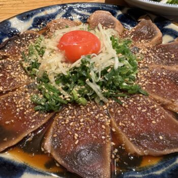 Thinly sliced seared tuna sashimi on a blue plate, sprinkled with sesame seeds and shredded onions and greens, topped with a cherry tomato.