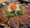 Thinly sliced seared tuna sashimi on a blue plate, sprinkled with sesame seeds and shredded onions and greens, topped with a cherry tomato.