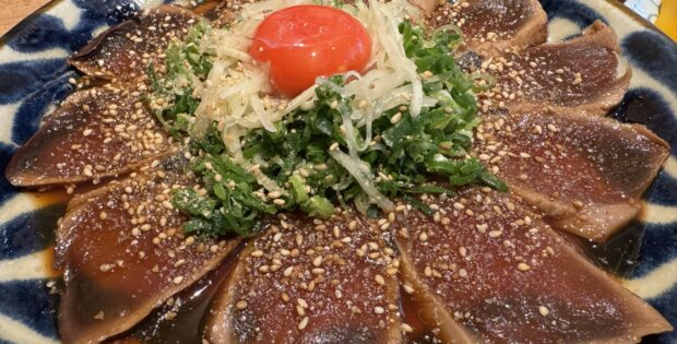 Thinly sliced seared tuna sashimi on a blue plate, sprinkled with sesame seeds and shredded onions and greens, topped with a cherry tomato.