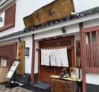 Traditional Japanese storefront with white walls, red wooden frames, white curtains, and a weathered wooden sign above the entrance.