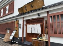 Traditional Japanese storefront with white walls, red wooden frames, white curtains, and a weathered wooden sign above the entrance.