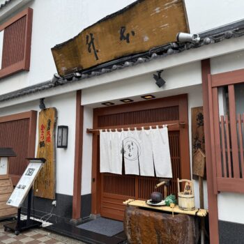 Traditional Japanese storefront with white walls, red wooden frames, white curtains, and a weathered wooden sign above the entrance.