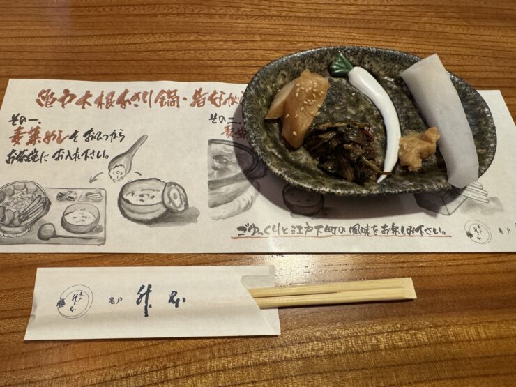 Ceramic plate with pickled vegetables topped with sesame seeds and a white fish cake strip, accompanied by chopsticks and printed menu sheets at a dining table.