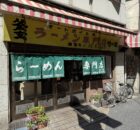 Restaurant storefront with a yellow sign, red-and-white striped awning, green text curtains, and bicycles parked outside.