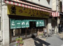 Restaurant storefront with a yellow sign, red-and-white striped awning, green text curtains, and bicycles parked outside.