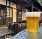 Plastic cup of pale beer with a foam head on a weathered blue table outdoors, rustic wooden building in the background.