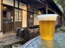 Plastic cup of pale beer with a foam head on a weathered blue table outdoors, rustic wooden building in the background.
