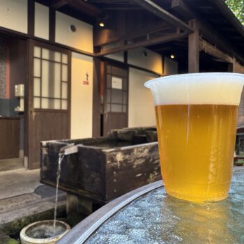 Plastic cup of pale beer with a foam head on a weathered blue table outdoors, rustic wooden building in the background.